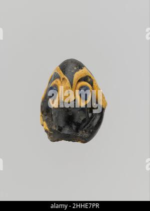 Glass pendant in the form of a demonic mask 6th–5th century B.C. Phoenician Semi-opaque deep yellow brown, appearing black, with additions in opaque yellow and translucent cobalt blue.Conical, splayed at bottom with rounded edge; rod hole underneath.Applied large arched eyebrows in yellow, eyes as blue knobs applied over large yellow circles, and yellow edge at bottom, extending round back edge.Large chip on front edge, suspension loop missing, and chip on face between eyes (perhaps nose missing); slight dulling and weathering, withencrustation inside rod hole.. Glass pendant in the form of a Stock Photo