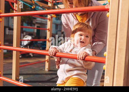 A little pretty child climbs the children's sports wall with the help of his mother. The concept of children's games and development. Stock Photo