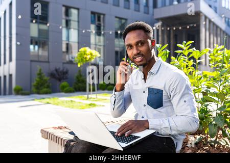 Successful African American businessman works with laptop and talks on the phone, looks at the camera and smiles rejoices outdoors, in casual clothes Stock Photo