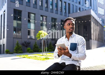 african american guy using tablet sitting on bench on city street on urban background. Black man reads or makes doing purchases holding a gadget or sm Stock Photo