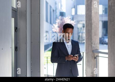 African American businessman grinds on video call uses phone and app, outside office, cheerful and successful man in business suit Stock Photo
