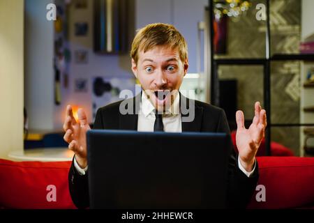 Surprised happy businessman wearing black suit using laptop amazed with open mouth for surprise. Stock Photo