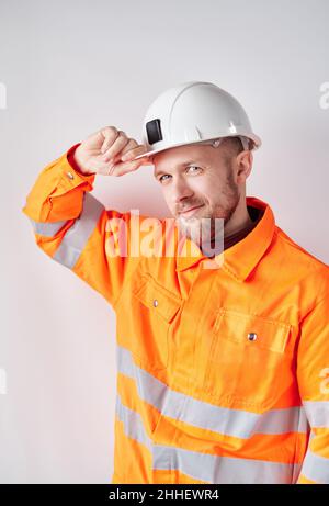 Building industry concept. Happy cute male caucasian technician, engineer, supervisor or building contractor portrait in white helmet and orange reflective jacket adjusting his working helmet Stock Photo