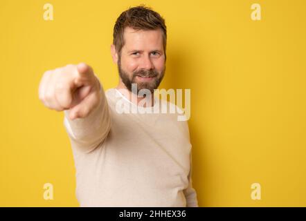 Smiling bearded man in casual sweater pointing finger at camera isolated on a yellow background Stock Photo