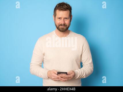 Young bearded man using smartphone standing isolated over blue background. Stock Photo