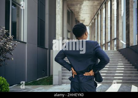 Businessman has severe back pain at work, african american freelancer man near office holding his back depressed trying to do massage Stock Photo