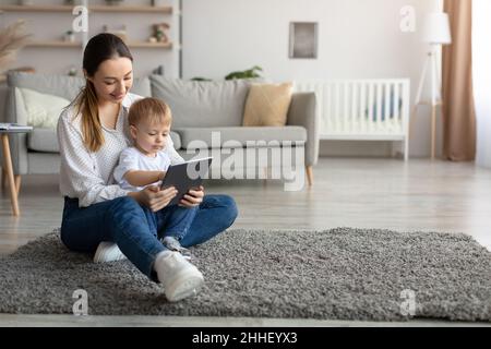 Happy mother and her kid son using digital tablet at home, watching development videos or surfing internet, copy space Stock Photo