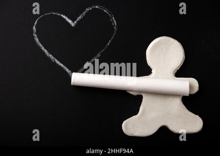 A small white plasticine man holding a chalk in his hands and drawing an outline of a white heart on a black board, copy space. Valentine's day concep Stock Photo