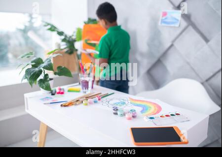Art supplies on table and behind boy standing near easel Stock Photo