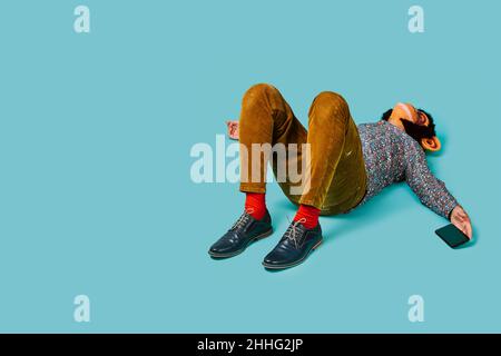 a fun man, wearing a monkey mask, is lying down on a blue background while is holding his smartphone in his hand, and some blank space on the left Stock Photo