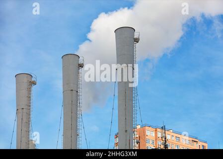 Three chimneys of a thermal power plant emitting steam under a clear blue sky against the backdrop of the top of a residential building. Stock Photo