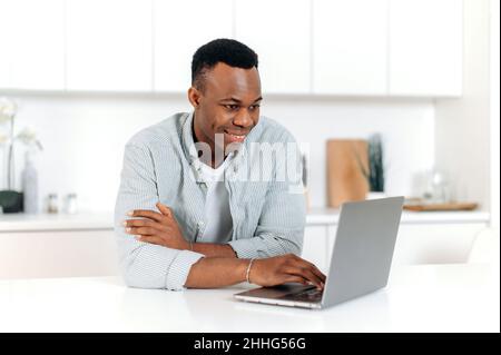 Pleasant modern black man, freelancer, designer or student, sitting at home in the kitchen, using laptop for studying or working on Internet improving his skills, smiling friendly. E-learning concept Stock Photo