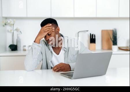 Tired dark-skinned guy in stress, freelancer, sitting at a table in kitchen, experiencing fatigue, headache, migraine, holding his head, closing his eyes, needs rest, overworked of computer work Stock Photo