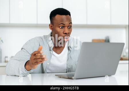 Shocked african american man. Goggled surprised black guy, sitting at a desk at home, looks at laptop in amazement, surprised, confused, received unexpected news, message, emotional face expression Stock Photo