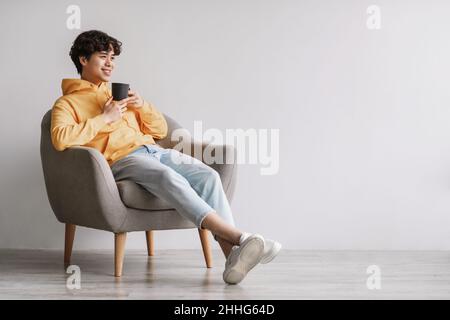 Full length of young Asian man drinking hot coffee in armchair against white studio wall, empty space Stock Photo
