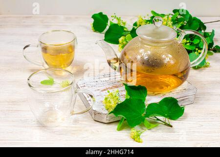 Empty glass teapot and two cups for tea on a wooden table against a background of lime leaves and flowers. Healing tea. Stock Photo
