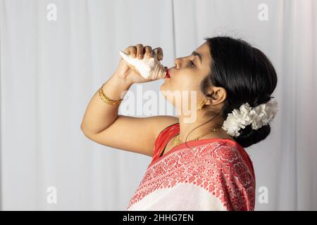 A beautiful Indian woman in red saree blowing conch shell on white background Stock Photo