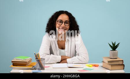 Happy young black female student sitting at desk with books and notebooks, smiling at camera on blue background Stock Photo