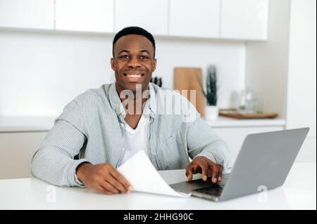 Positive pleasant african american guy, student or freelancer, in stylish casual clothes, sits with laptop at the kitchen at home, looking at the camera, smiling friendly Stock Photo