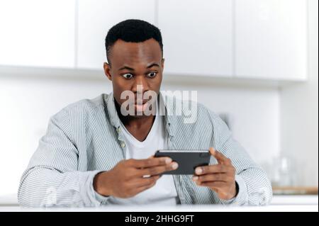 Stunned shocked frightened African American man, sit at a table in a kitchen, looks at the phone screen in amazement, receives unexpected news, wins or big profits, surprised puzzled facial expression Stock Photo