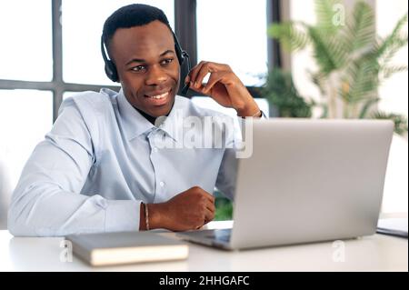 Successful pleasant African American guy in headphones, hotline operator, support worker, online consultant is sitting at a laptop in the office, talking to a client on a video call, smiling friendly Stock Photo
