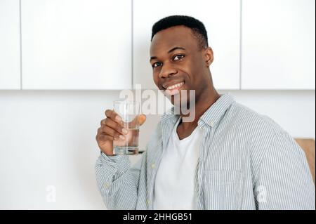 Healthcare concept. Close-up positive african american young man, in casual wear, stands at home in the kitchen, holds a glass of pure water, drinks a daily amount of water, takes care of health,smile Stock Photo