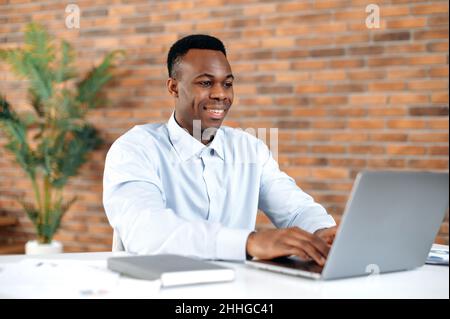 Positive confident black man using laptop in modern office. Successful businessman, freelancer, manager, in formal shirt, browsing email, working and typing on laptop, working on a project, smiling Stock Photo