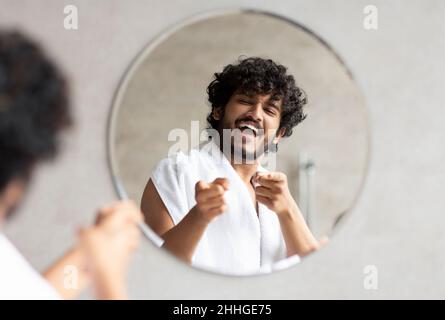 Excited indian guy pointing at mirror in bathroom with two fingers, standing with towel on shoulder Stock Photo