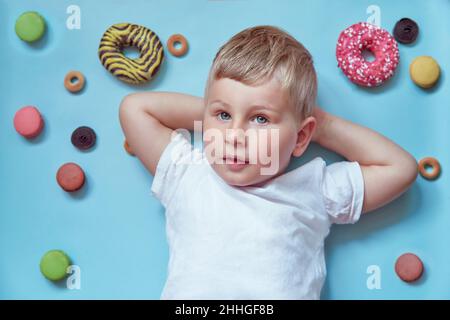 Cute smiling child on donuts and french macarons on blue background. Mock up white T-shirt. National Donut Day concept. Happy childhood concept