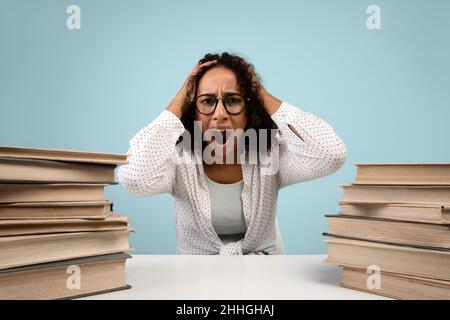 Desperate black female student holding her head and screaming, surrounded by books on blue studio background Stock Photo