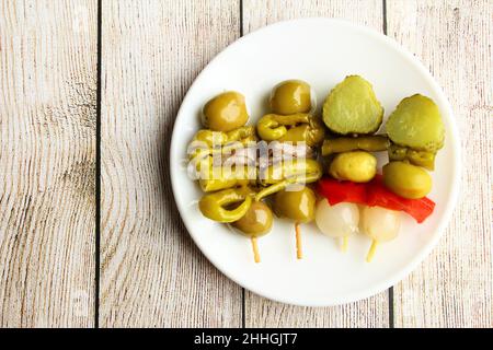 Pickled skewers of olives, anchovy fish, sweet chili pepper, cucumber on white plate photo isolate on clear wooden background top view copy space Stock Photo