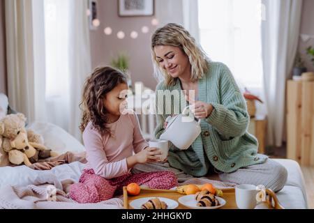 Happy mother with her little daughter having breakfast together in bed at home. Stock Photo