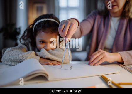 Mother helping her daughter with homework, drawing a circle with comasses in evening at home. Stock Photo