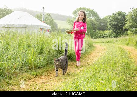 Girl in pajamas walking in grassy field with cat near yurts in campsite Stock Photo
