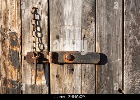 Wooden barn door with metal hasp lock held in place by a metal gravity  pin on a chain, in bright light with strong shadows Stock Photo