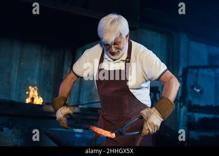 Focused caucasian craftsman in protective apron and gloves forging steel on anvil. Mature blacksmith using industrial tools for work with molten metal. Stock Photo