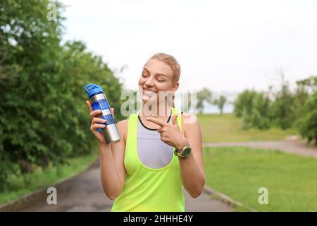 Young smiling woman in sportswear pointing at bottle of protein shake outdoors Stock Photo