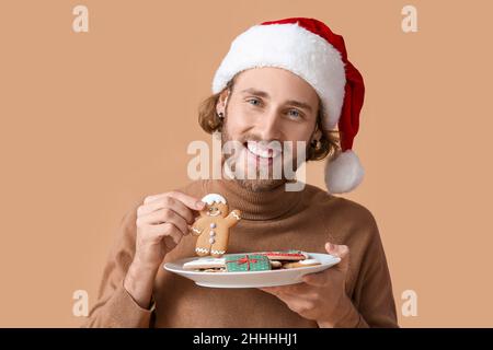 Happy young man in Santa hat holding plate with gingerbread cookies on beige background, closeup Stock Photo