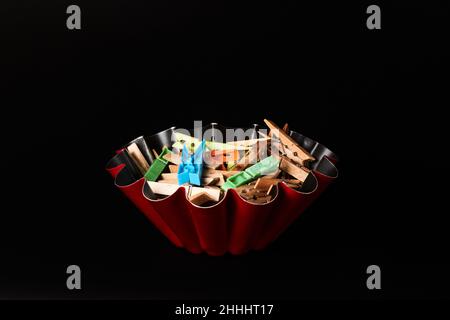 Red flan box on a black background filled with clothespins, some made of wood and others made of colored plastic. Copy space. Horizontal photography. Stock Photo