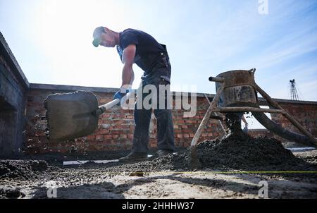 Male builder in work clothes, standing on construction site and distributing working material by shovel onto base of future floor. Repair of floor covering outside. Stock Photo
