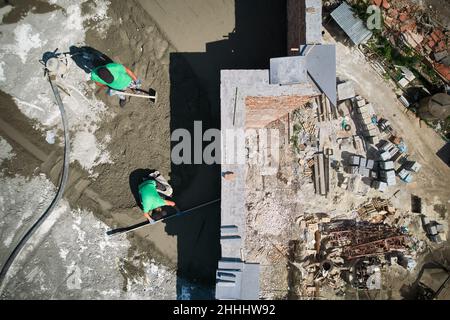 Two men builders screeding floor at construction site. Top view of male worker standing near concrete screed mixer machine and holding shovel while colleague using leveling surface with straight edge. Stock Photo