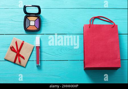 Red lipstick, eye shadows, paper bag and gift box on blue wooden background. Fashion accessories. Top view and mock up. Stock Photo