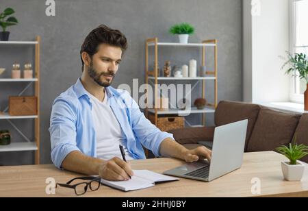 Young man sitting at desk with laptop computer, having online class and taking notes Stock Photo