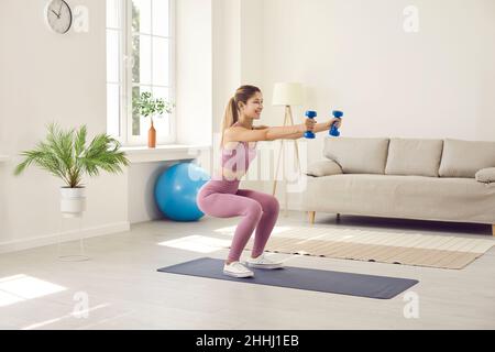 Beautiful and slender young woman performs sports exercises at home using dumbbells. Stock Photo