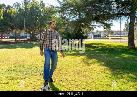 Hispanic man in his 40s playing soccer on the grass of a nice park wearing jeans and a shirt. Fun concept Stock Photo