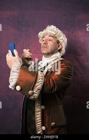 Serious man in classical renaissance clothes using an old monocle to see the mobile phone screen. Stock Photo