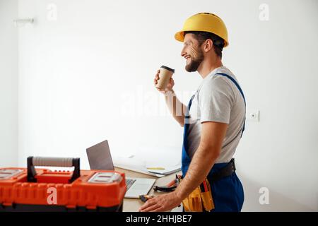 Side view of smiling handyman in uniform and hard hat drinking coffee during a break while doing renovation work indoors Stock Photo