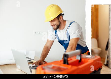 Smiling repairman in uniform and hard hat using laptop, looking through details of an order, preparing for renovation work Stock Photo