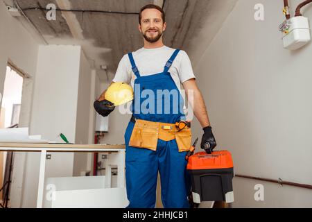 Hard working contractor, handyman in uniform with toolbox smiling at camera while standing indoors Stock Photo