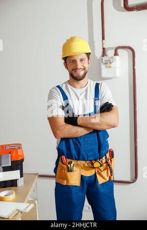 Professional electrician technician in uniform, hard hat and protective gloves smiling at camera while standing with arms crossed indoors Stock Photo